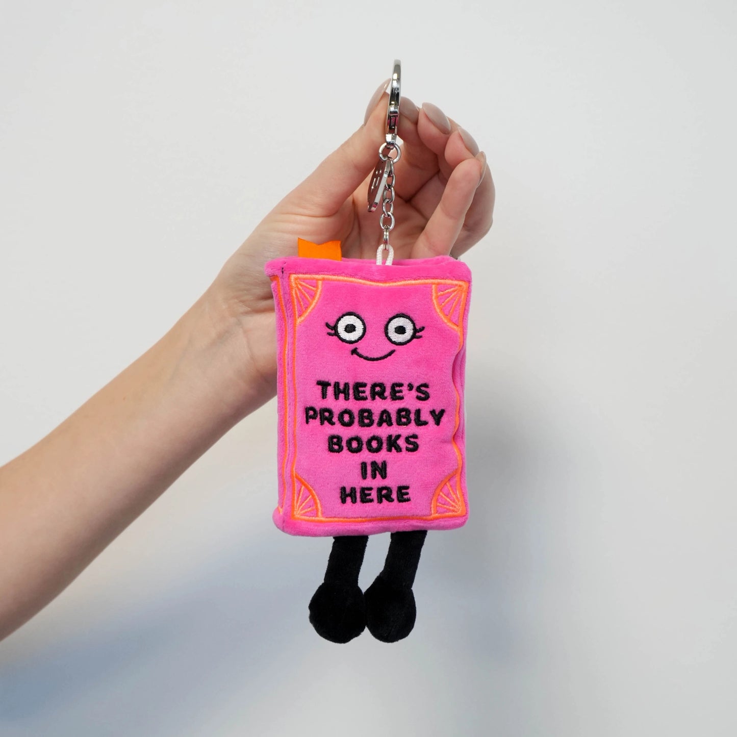 Pink book plush bag charm held in hand showing detailed embroidery, dangling legs, and silver keychain ring with bookmark detail
