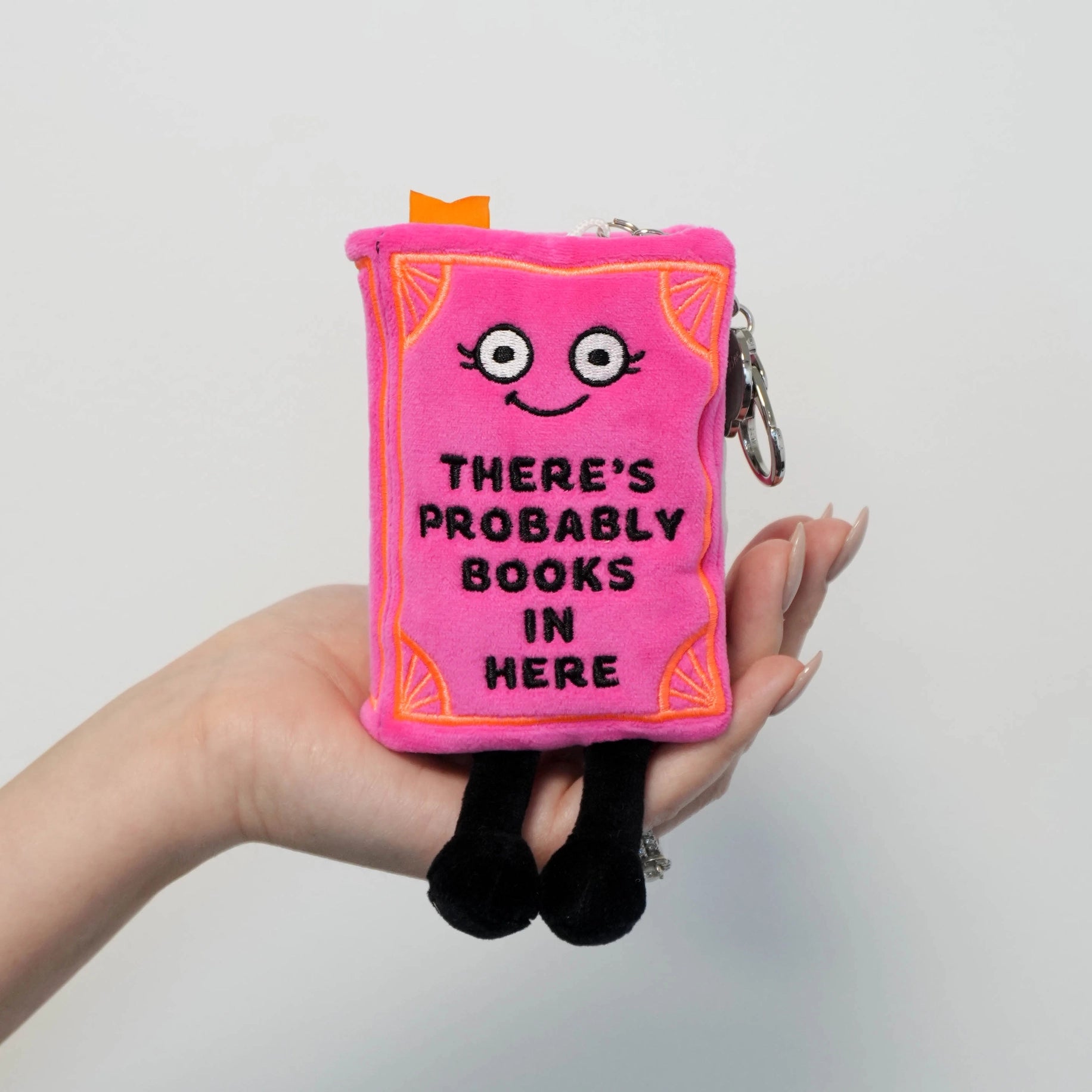 Hand holding pink book plush bag charm with orange embroidered corners, dangling black legs, and lobster-clasp keychain hardware