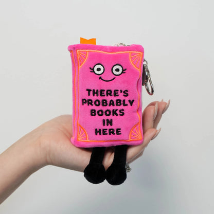 Hand holding pink book plush bag charm with orange embroidered corners, dangling black legs, and lobster-clasp keychain hardware