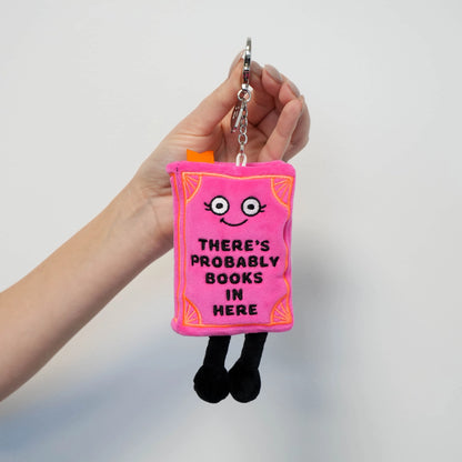 Pink book plush bag charm held in hand showing detailed embroidery, dangling legs, and silver keychain ring with bookmark detail