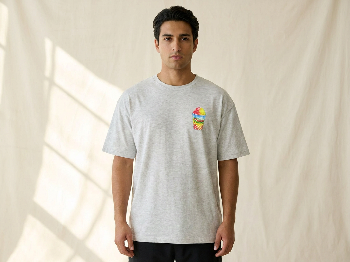 A man wearing a light gray heather crewneck t-shirt with a small colorful water ice graphic on the left chest, standing against a draped beige fabric backdrop.