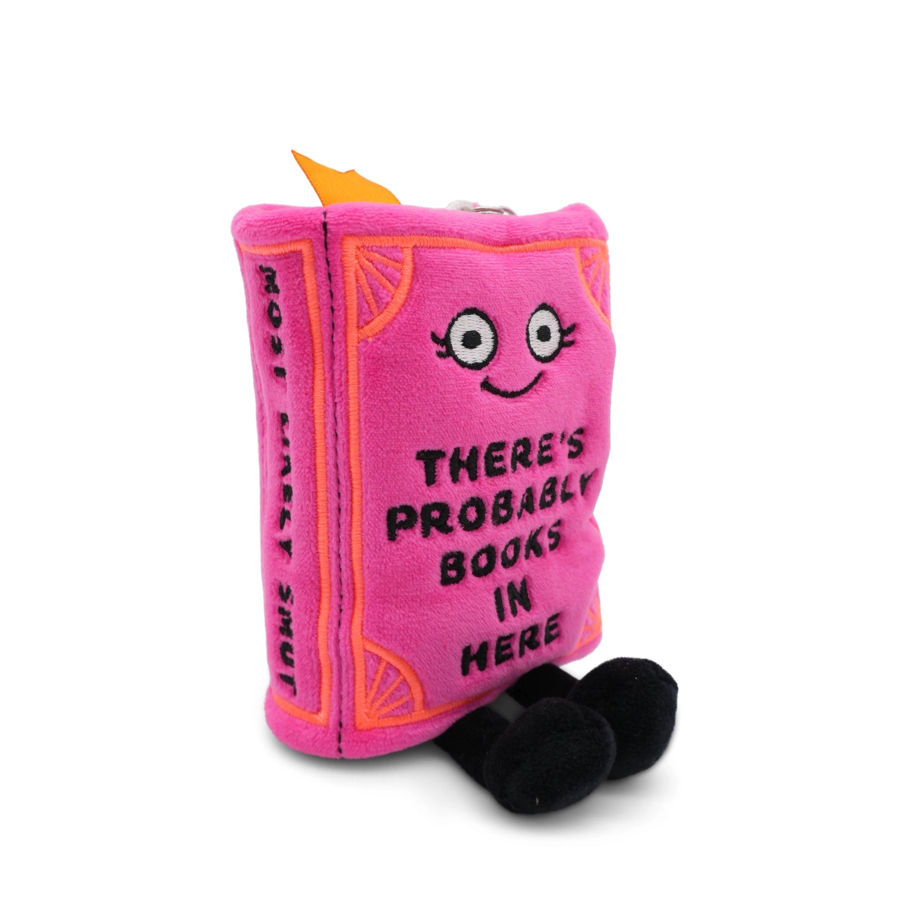 Pink book plush bag charm standing upright showing embroidered smiling face on cover and "MOST LIKELY SMUT" text running vertically along the spine